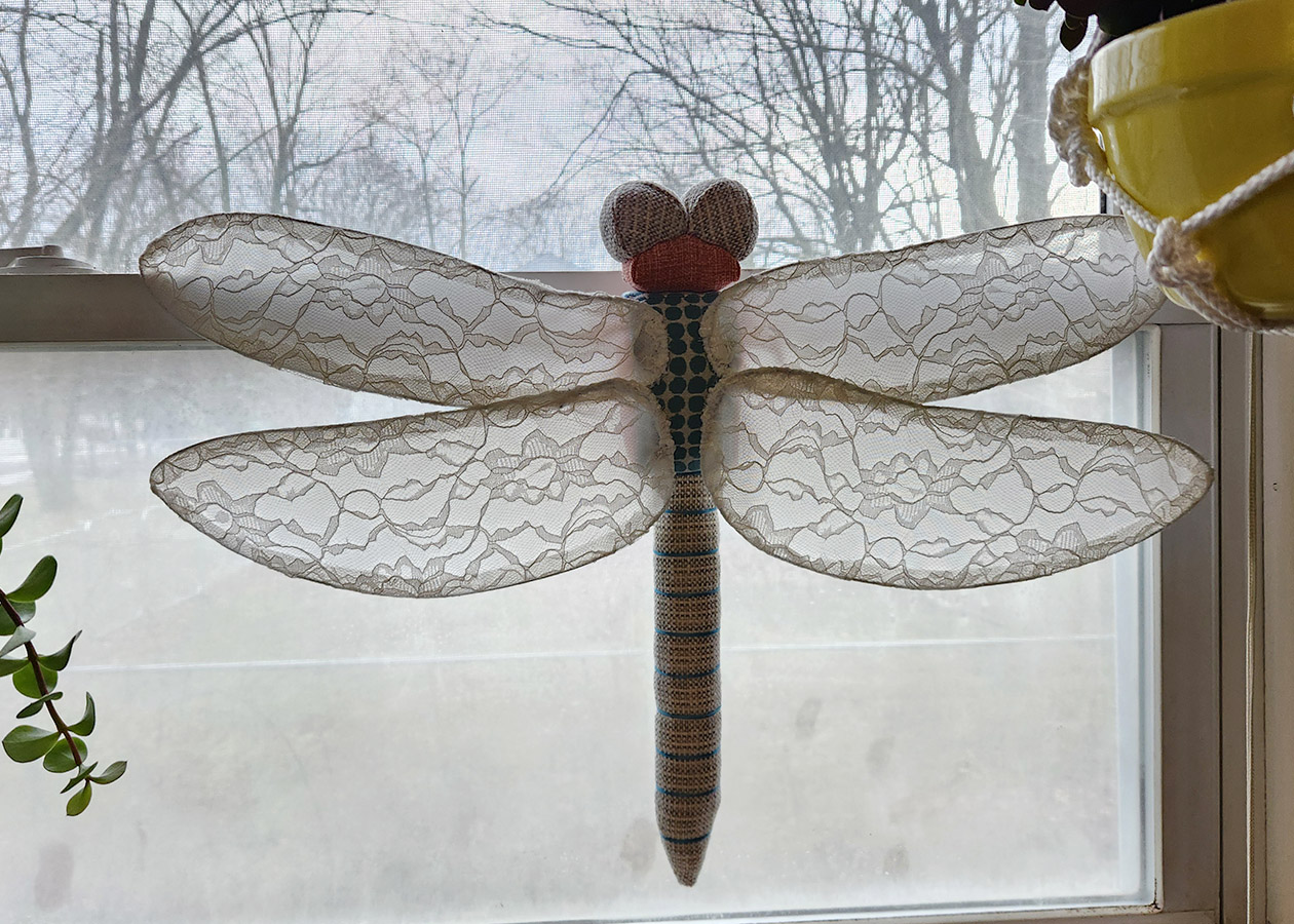 dragonfly fiber art sculpture with lace fabric wings perched on a window