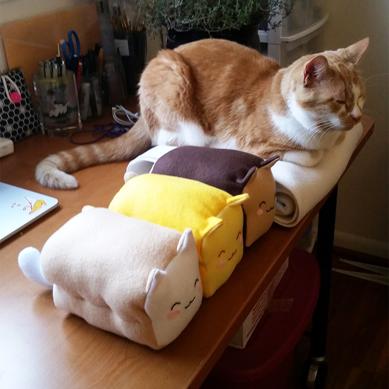 3 Meow-Bao plushies sitting with an orange and white cat loafing