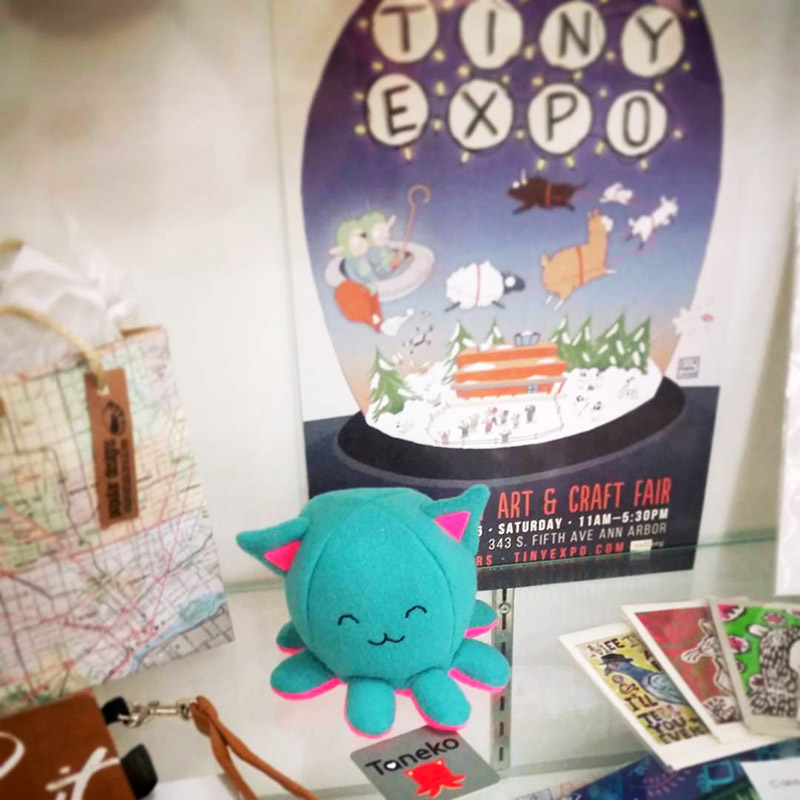 Taneko plushie in front of Tiny Expo event poster