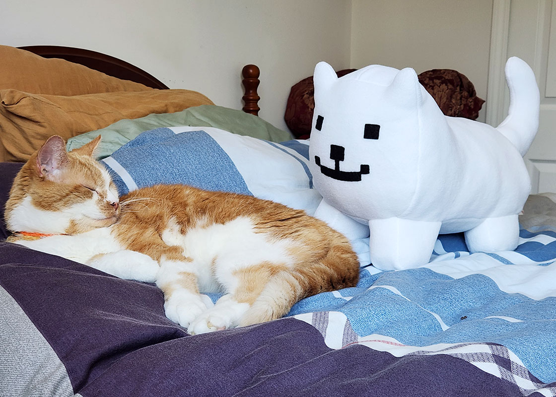 Undertale Annoying Dog plush toy on a bed with orange and white tabby cat