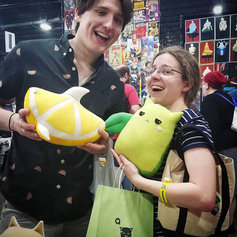 two people smiling holding plushies of a Lemon Shark and Cactus Cat at Motor City Comic Con